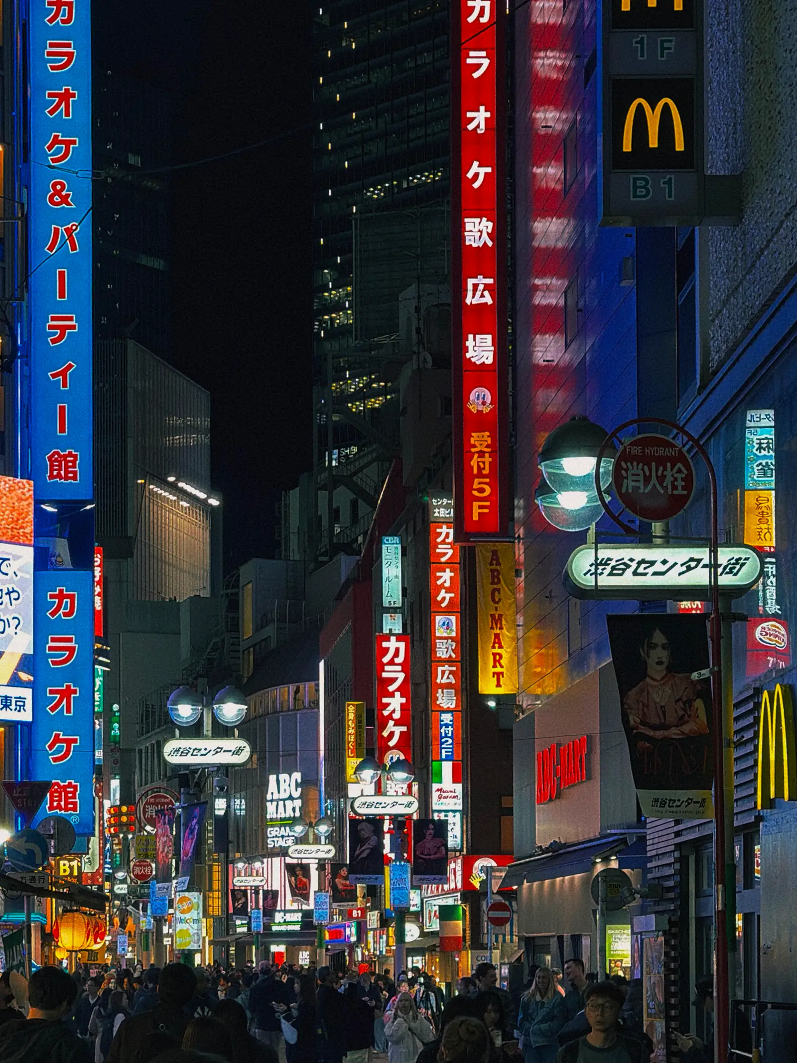Busy nighttime street in Tokyo filled with pedestrians, glowing neon signs, karaoke advertisements, and illuminated storefronts.