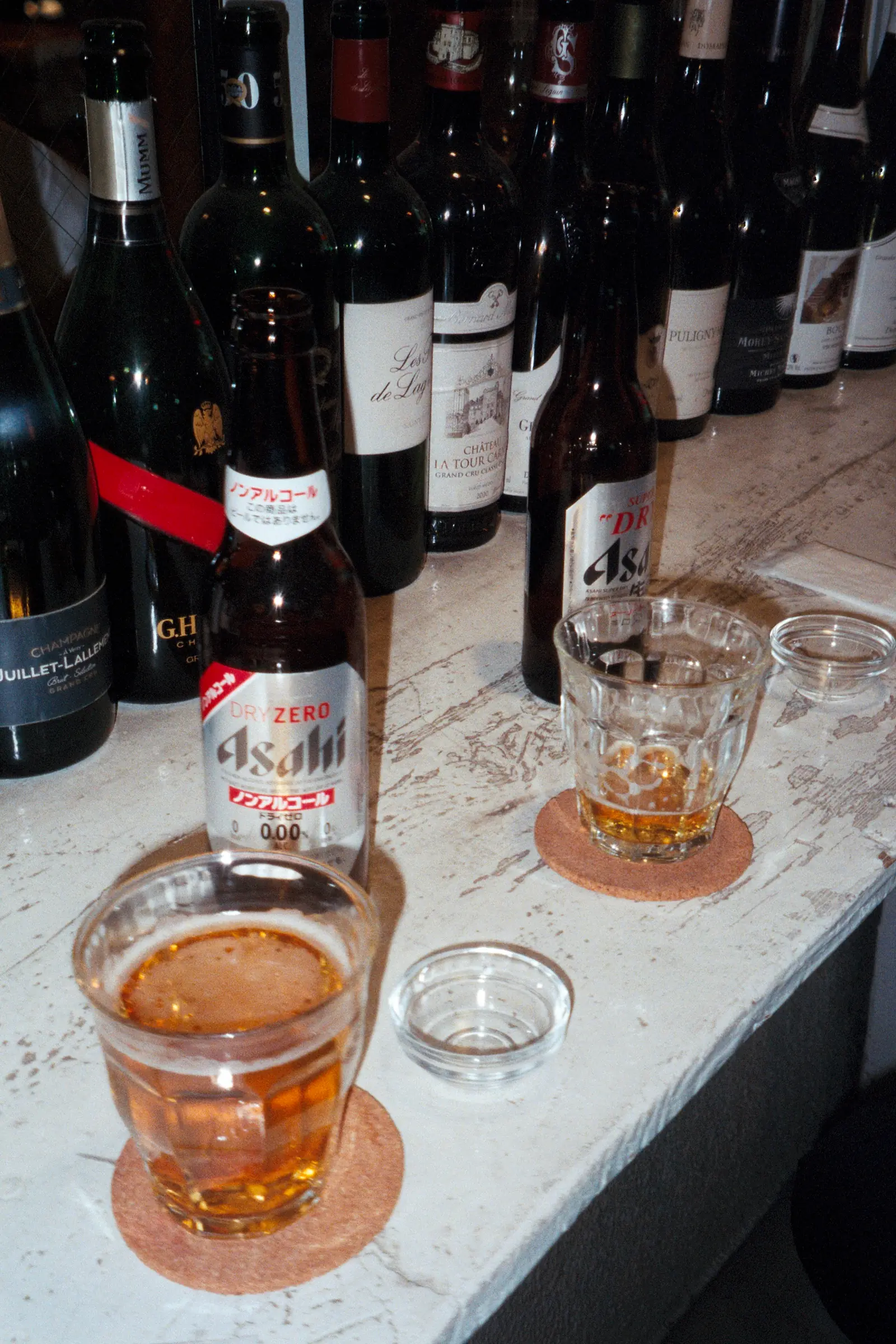 Bottles of wine and non-alcoholic Asahi beer lined up on a worn countertop beside two partially filled glasses in a dimly lit bar setting.