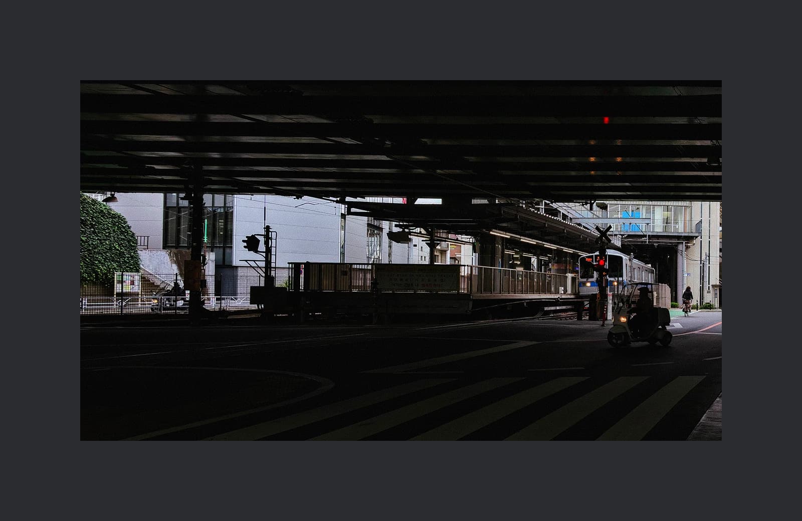Dark urban train platform beneath an overpass with a train arriving and a small vehicle waiting to cross the tracks.