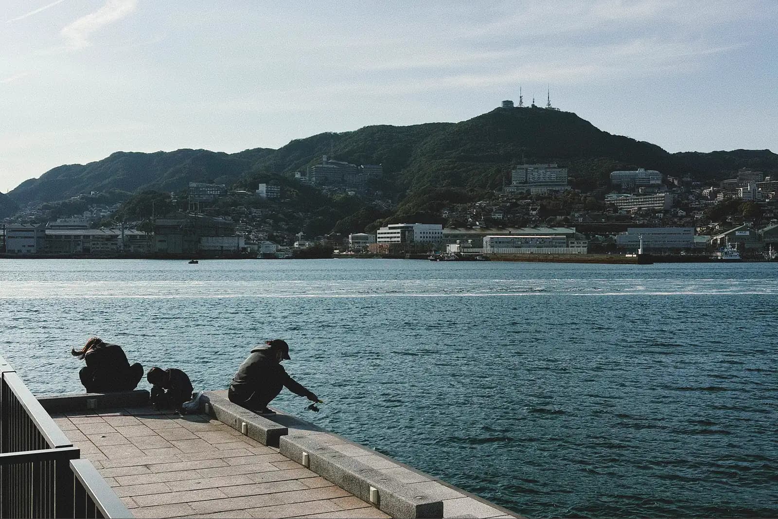 Three people sitting by the Nagasaki waterfront, with one person fishing while looking out across the water toward green hills and buildings under soft afternoon light
