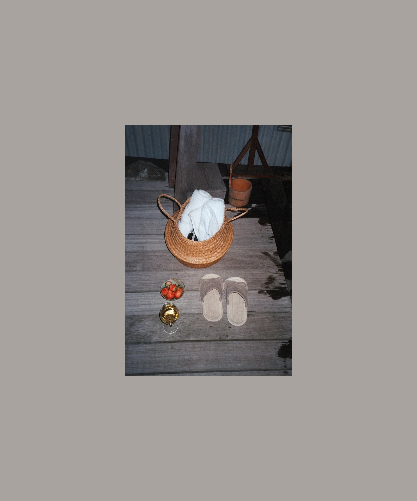 Straw basket, slippers, strawberries, and a glass of white wine arranged on a wooden deck at an onsen in a quiet nighttime setting.