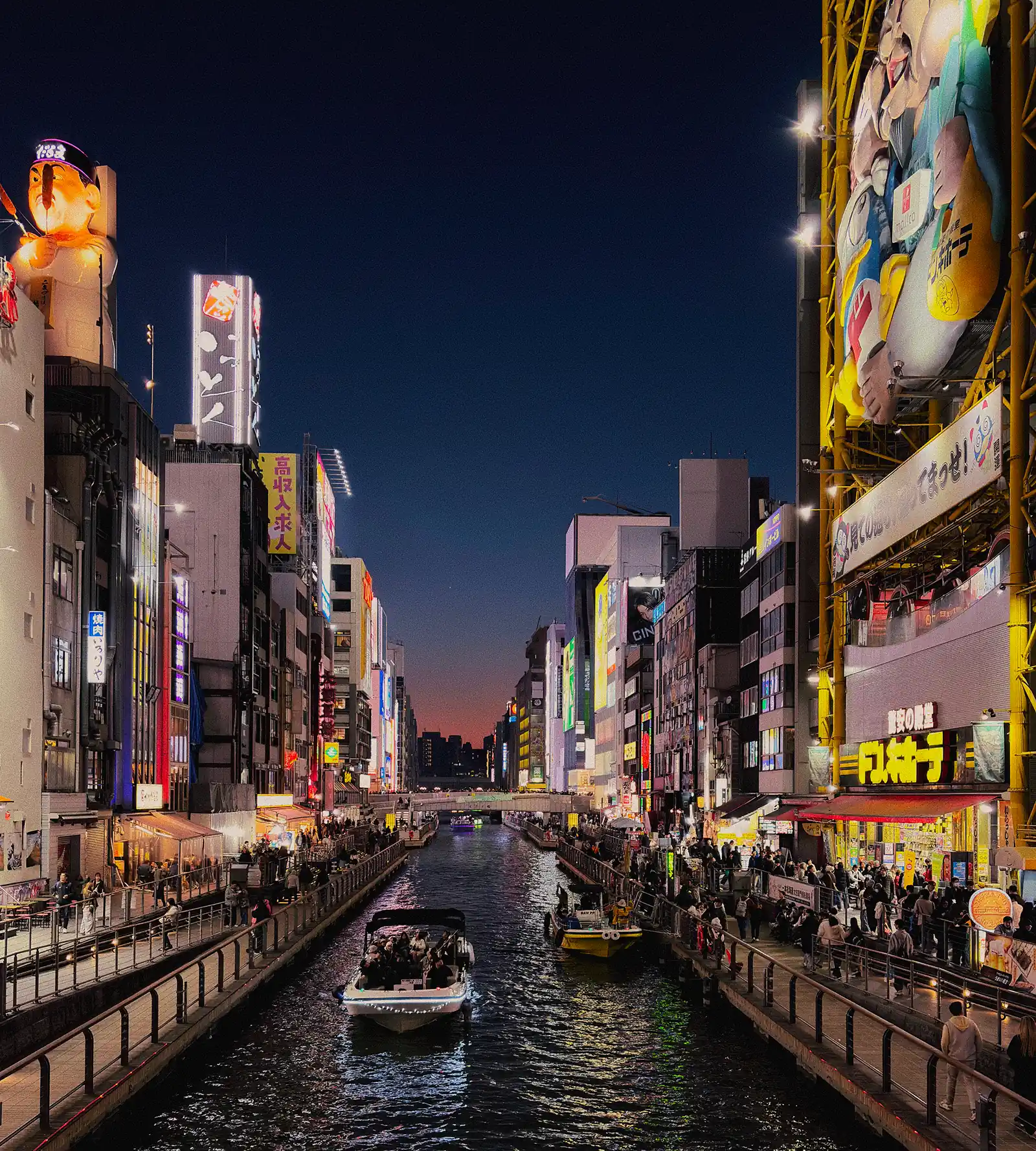 Illuminated canal street in Osaka at dusk with boats on the water, crowds along the promenade, and colorful billboards glowing against the evening sky.