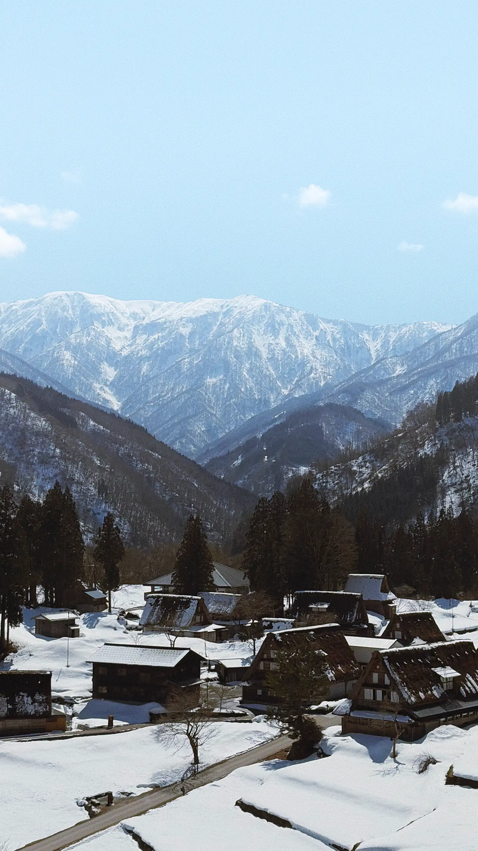 Snow-covered village in Gokayama with traditional houses in the foreground and large mountain peaks under a pale blue sky.