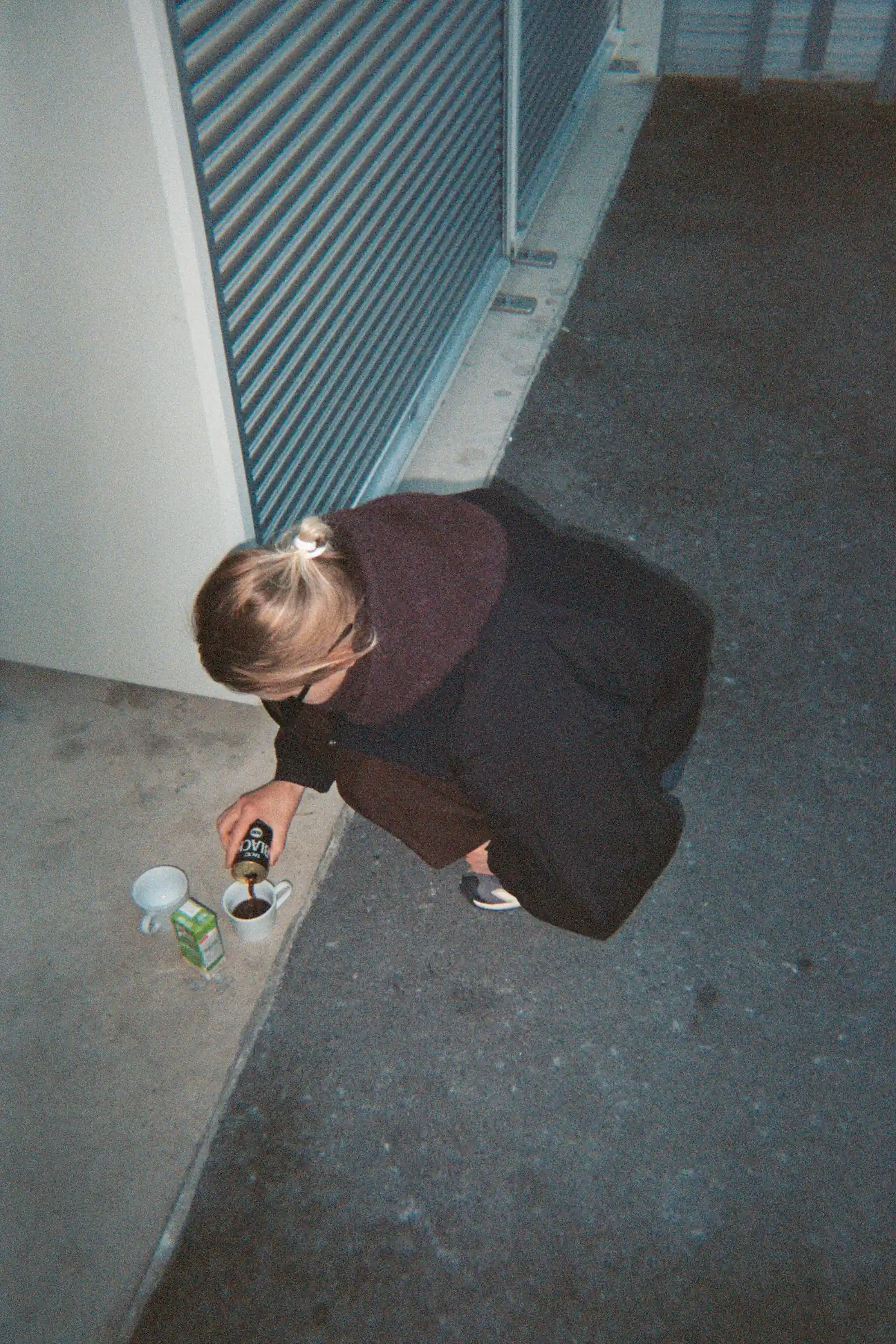 Person crouching outdoors early in the morning while pouring coffee into a mug beside a building.