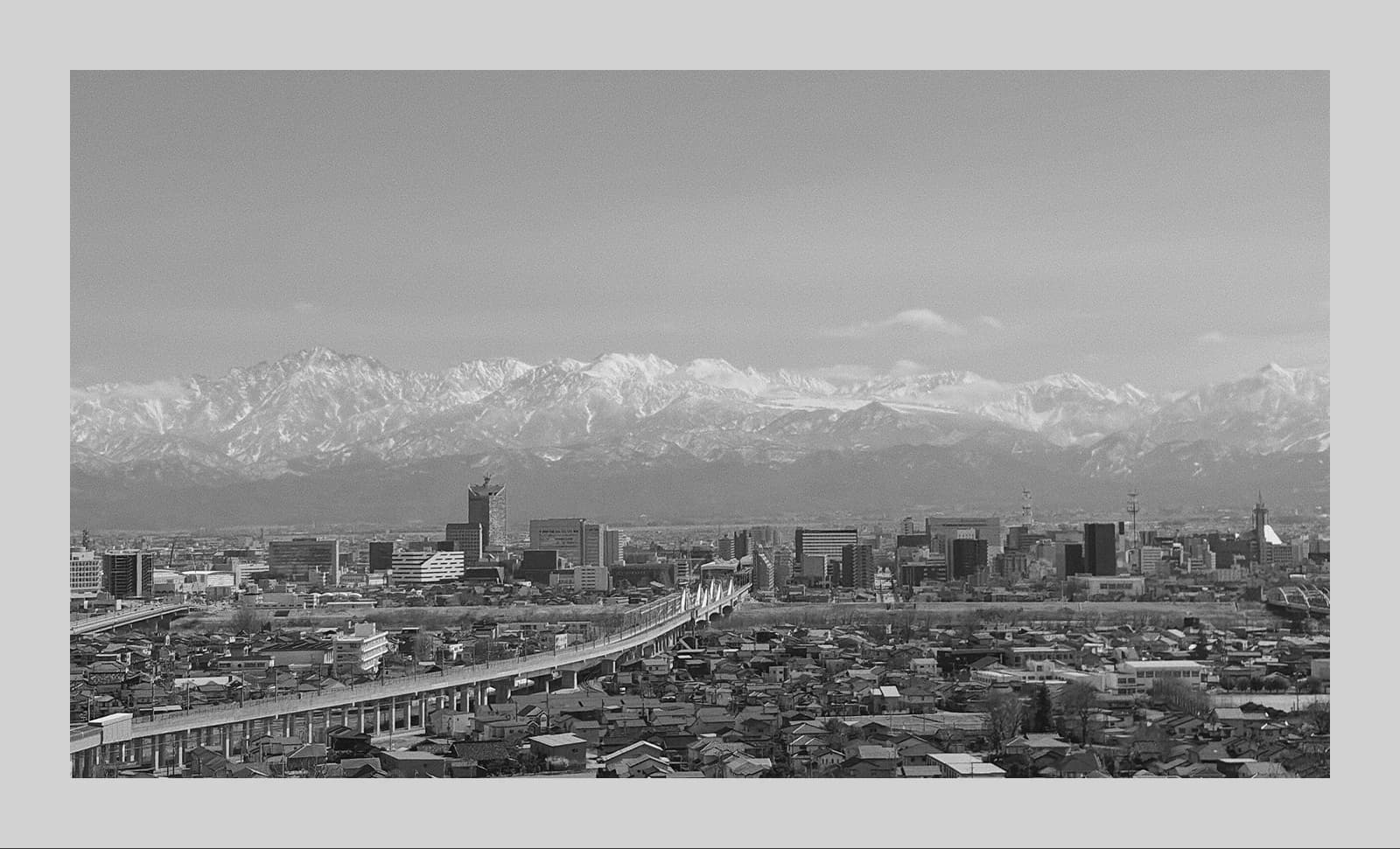 Black-and-white panoramic view of Toyama with elevated rail tracks in the foreground and snow-covered mountains in the distance.